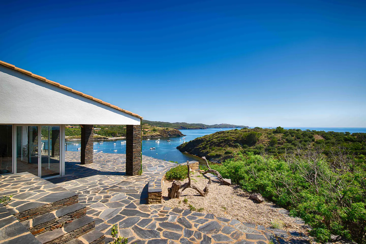 Stone patio of a modern home overlooking a scenic blue bay with boats, surrounded by lush green hills under a clear sky.
