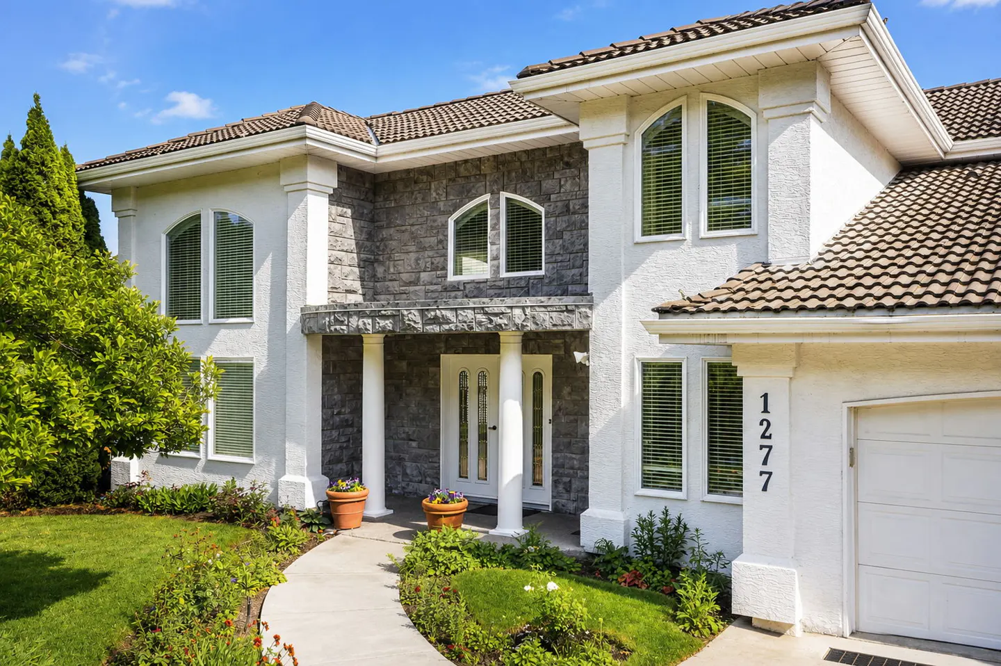 Two-story home with white stucco, grey stone accents, arched windows, a white front door, and a concrete walkway.