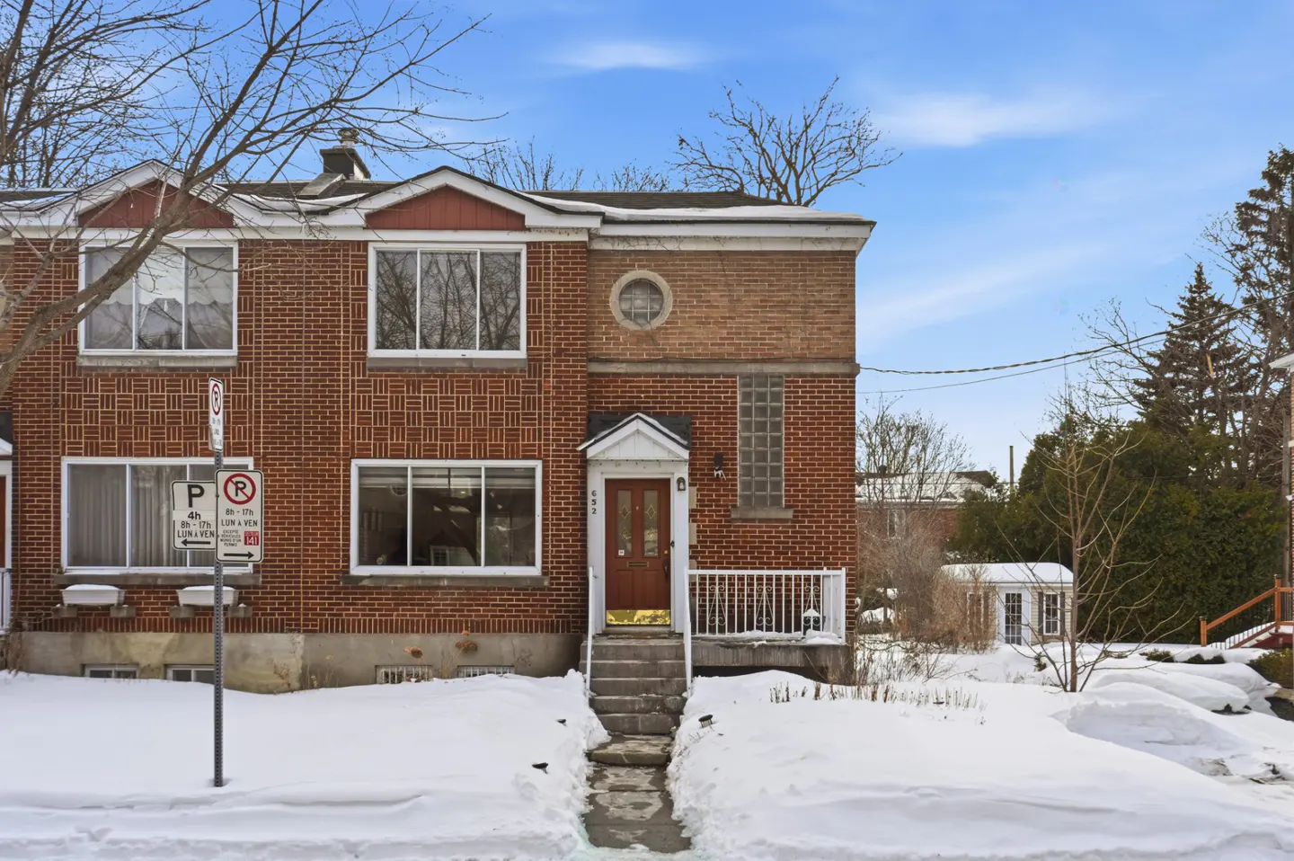 Red brick townhouse with a brown door and concrete steps, surrounded by snow on a sunny winter day.