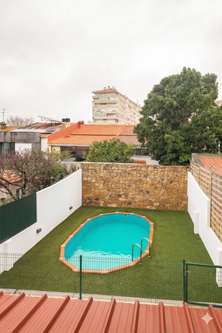 Aerial view of a backyard with a turquoise pool, green lawn, stone wall, and white fences, under an overcast sky.