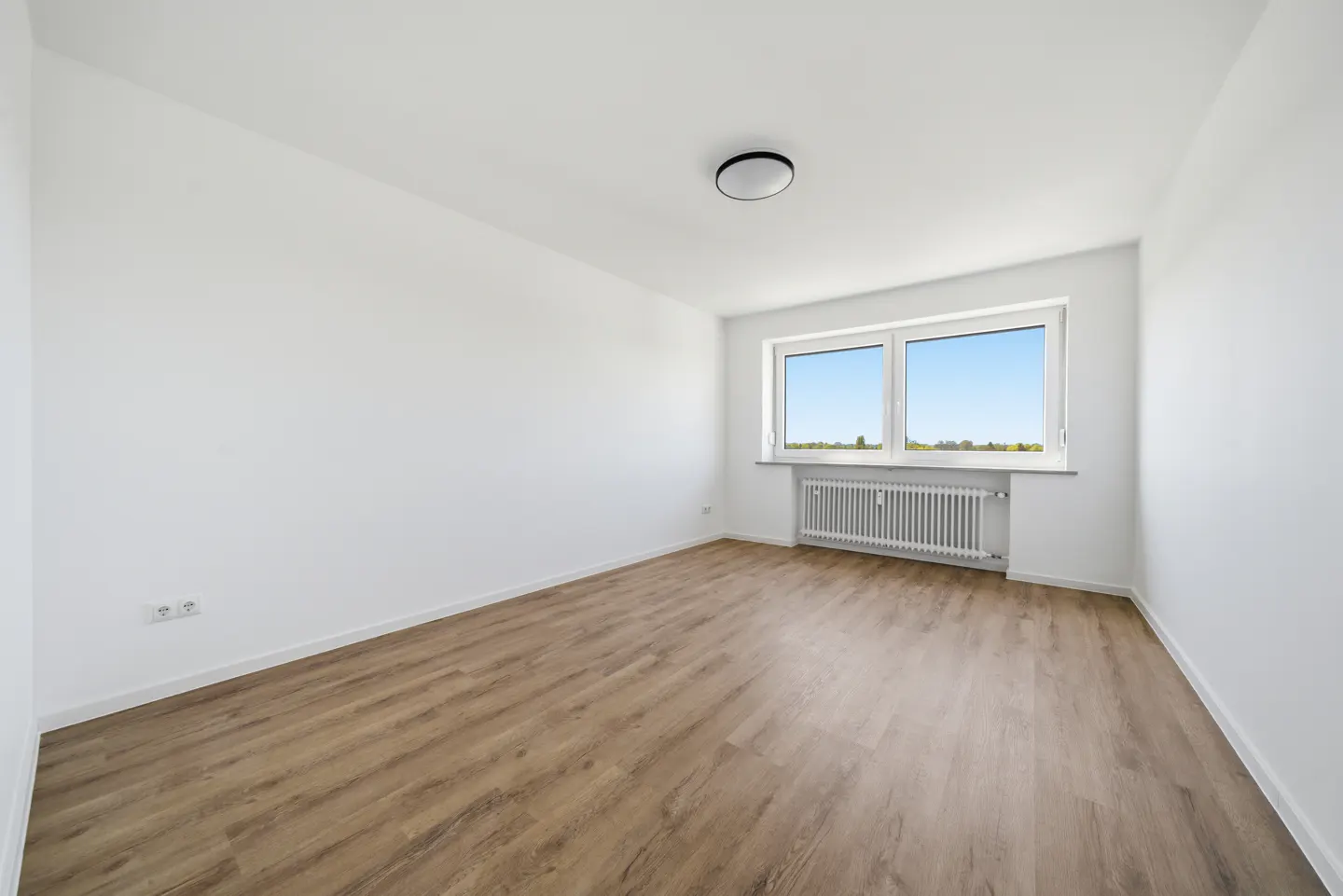 Bright, empty room with wood-look floors, white walls, and a window showing a blue sky. A white radiator sits beneath the window.
