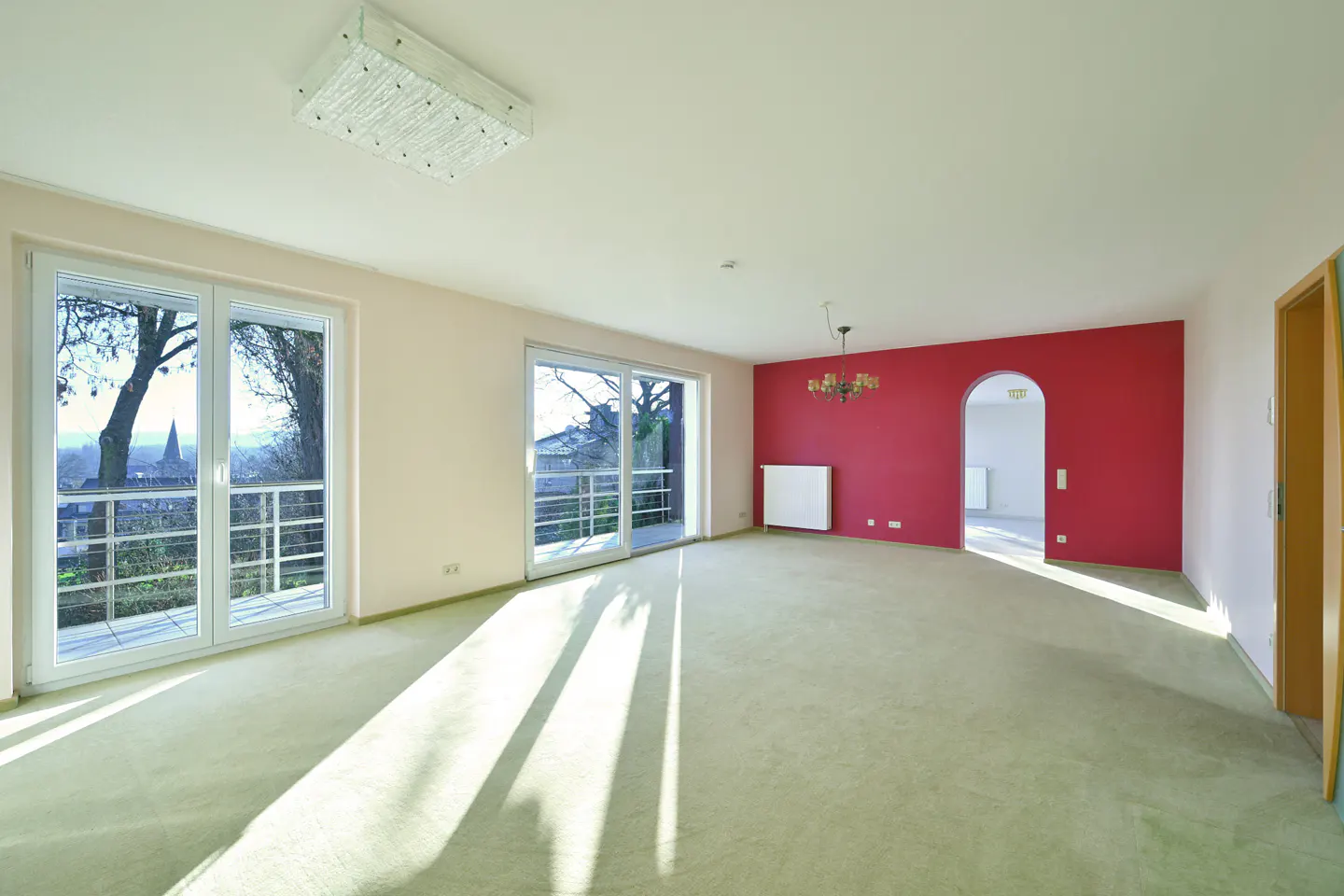 Bright, empty room with green carpet, white walls, and a red accent wall. Large windows let in sunlight. A chandelier hangs near an arched doorway.