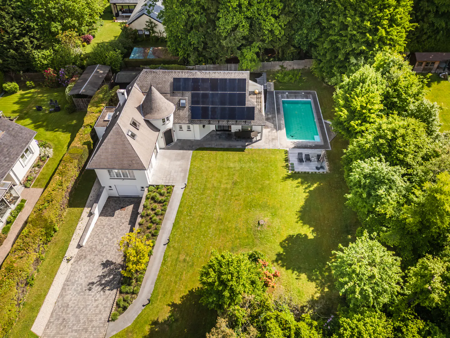 Aerial view of a white house with a pool, solar panels, and a large green lawn surrounded by trees.