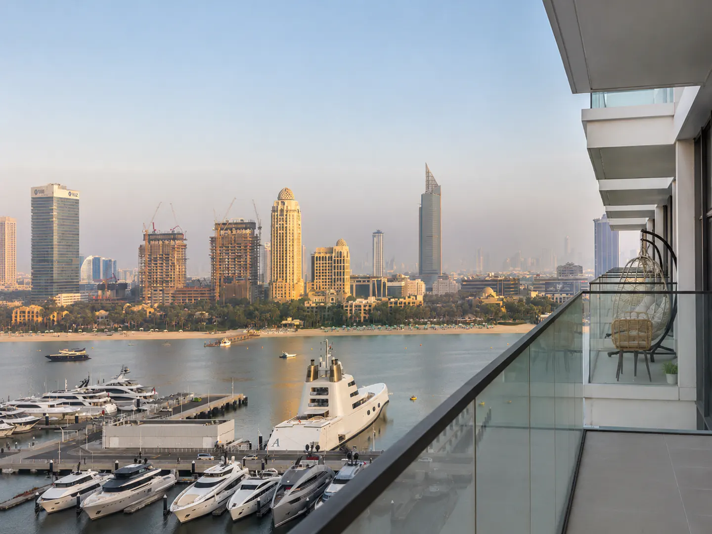Balcony view of Dubai skyline, yachts, and ocean. A hanging chair sits on the balcony with glass railings.