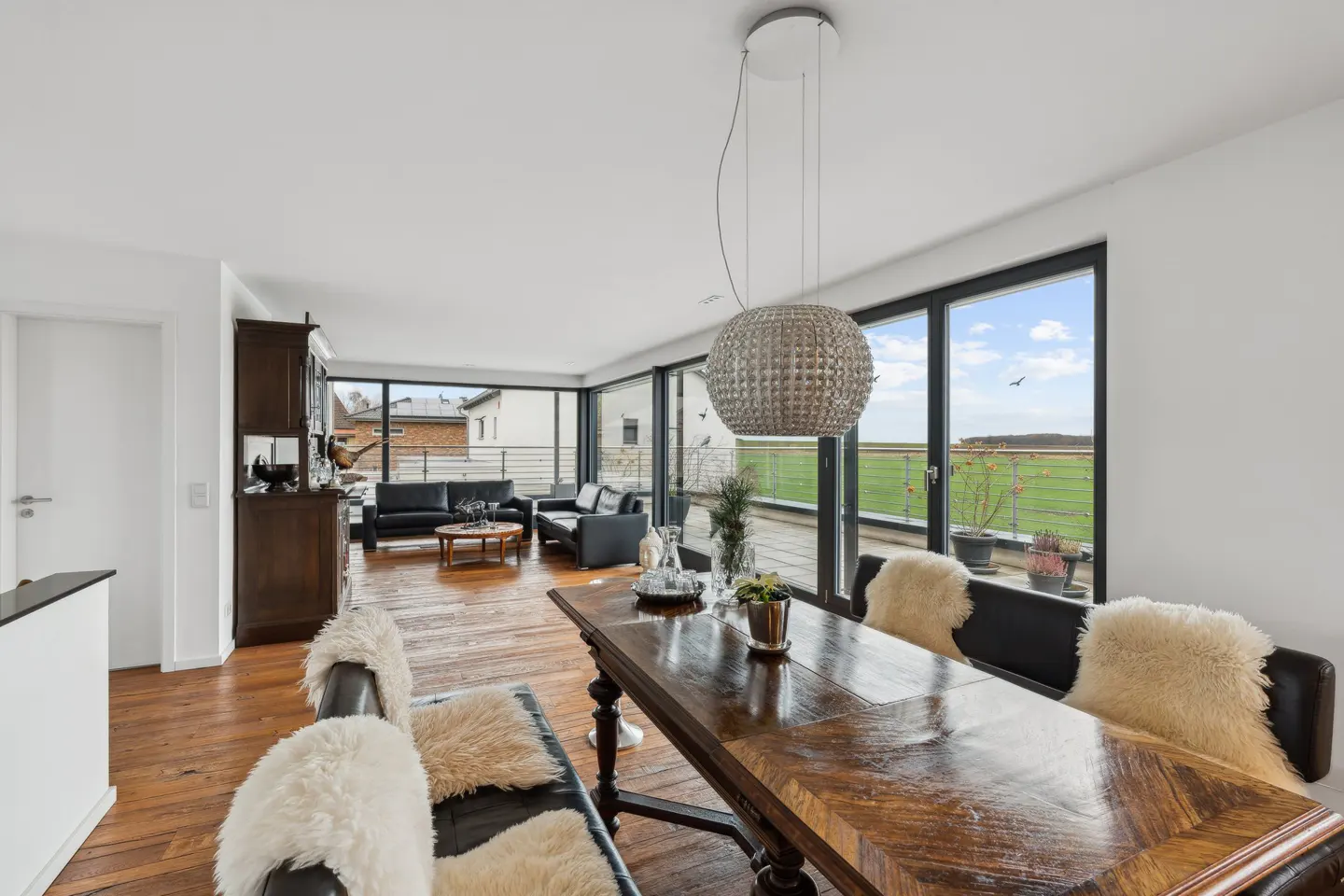 Bright living room with wood floors, a dining table with fur throws, and a view of a green field through large windows.