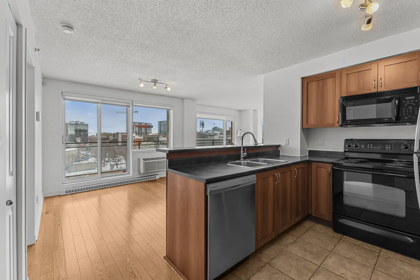 Open-concept kitchen with wood cabinets, black appliances, and a stainless steel dishwasher. Large windows offer city views.