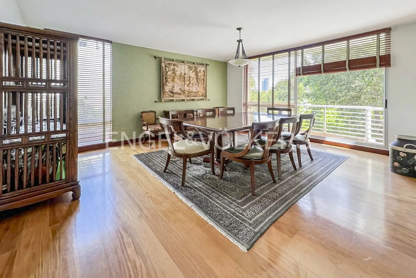A dining room with a large wooden table, chairs, and a rug on a hardwood floor. A tapestry hangs on the green patterned wall.