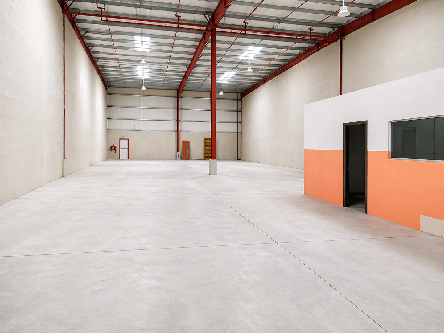 A large, empty warehouse with a concrete floor, red steel beams, and a white and orange office space.