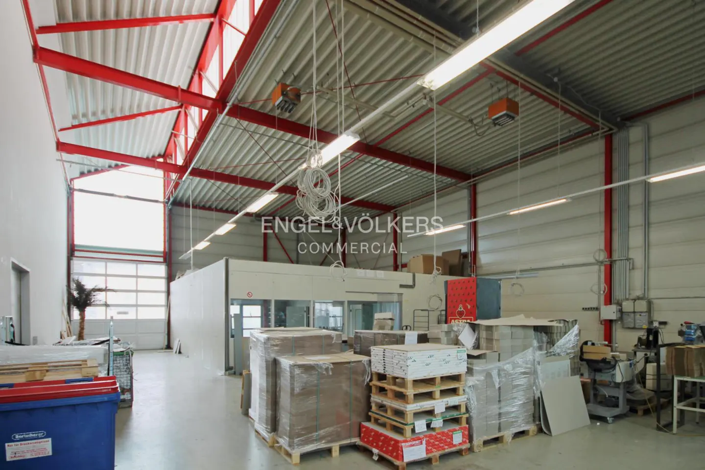 A bright warehouse interior with red steel beams, pallets of goods, and a white office structure.
