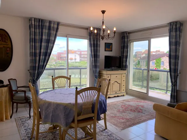 Bright dining room with a round table, wooden chairs, and blue plaid curtains framing a balcony view. A chandelier hangs above.