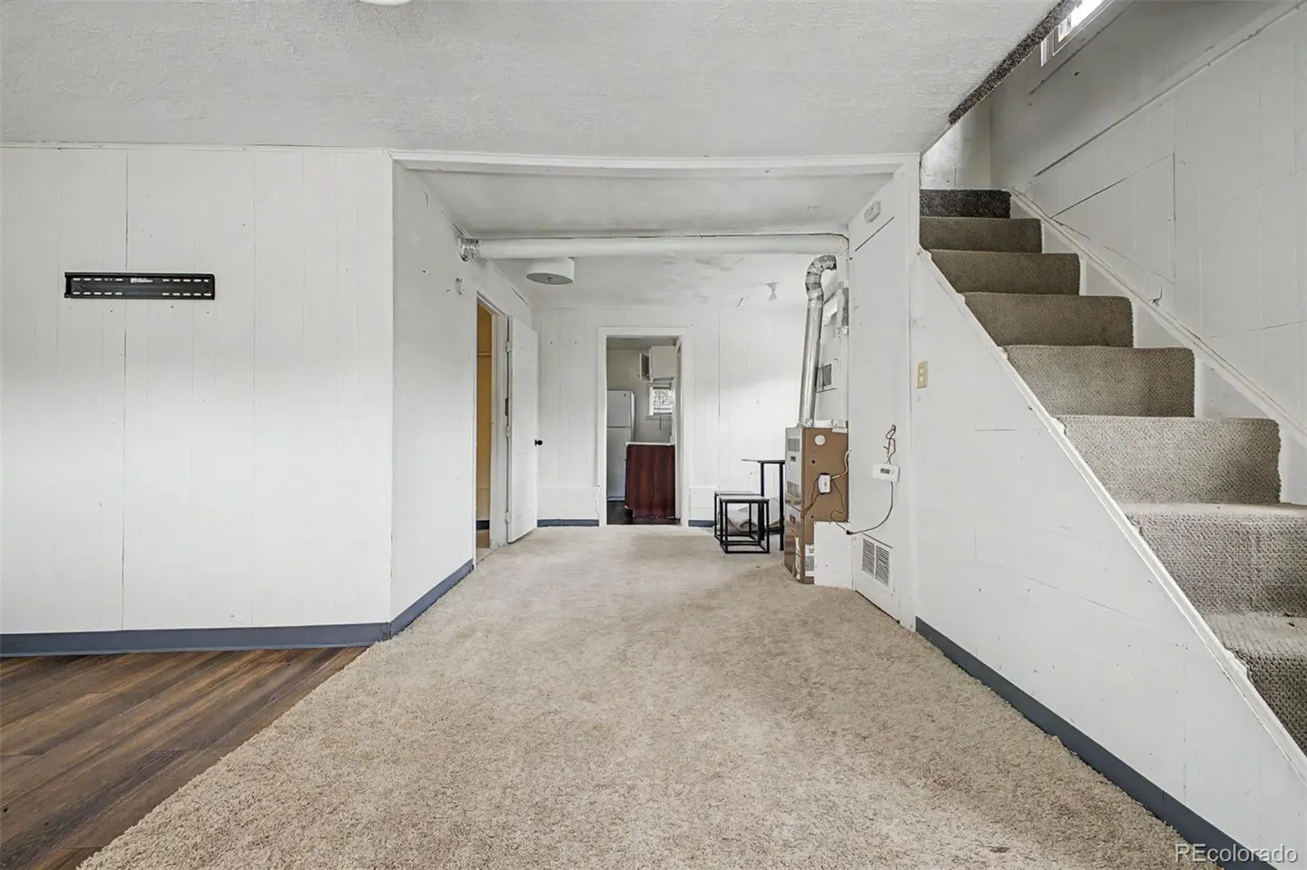 Unfinished basement with beige carpet, white paneled walls, a staircase on the right, and a view into a small kitchen.