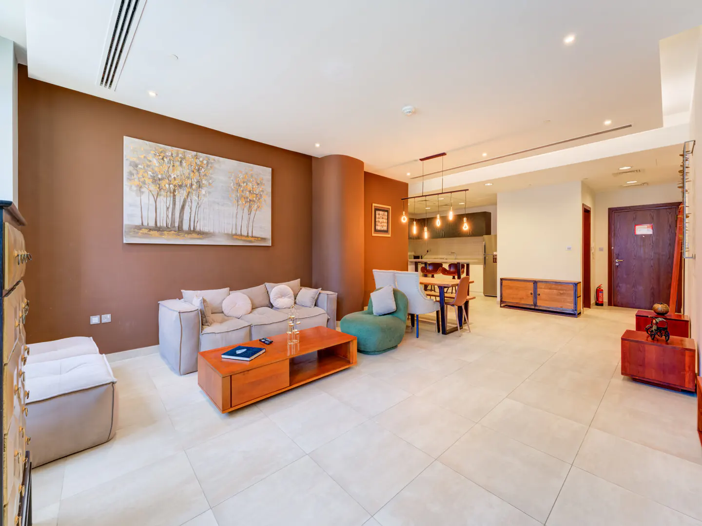A modern living room with a brown accent wall, a gray sofa, and a wooden coffee table. A dining area and kitchen are visible in the background.