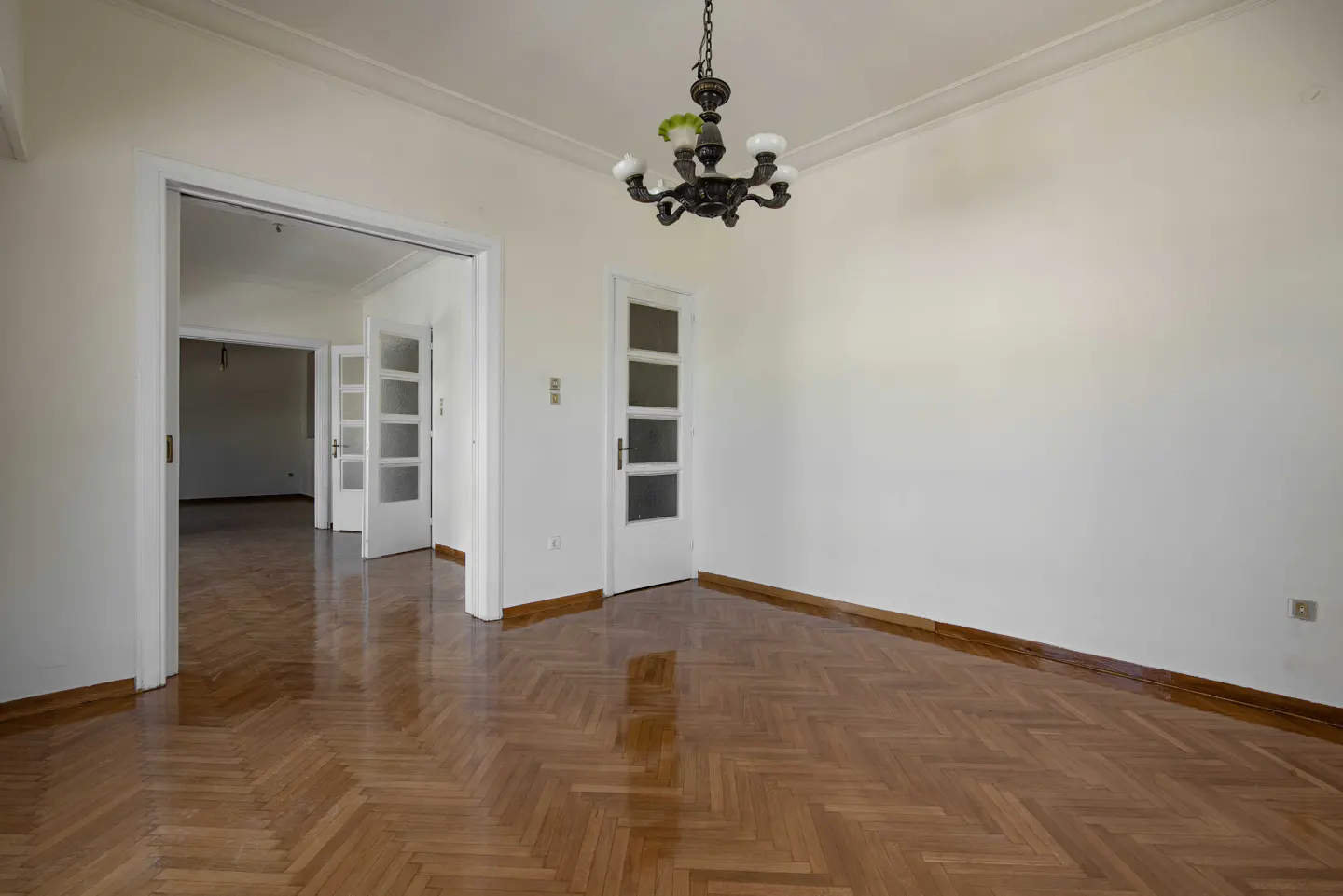Empty room with herringbone wood floors, white walls, and a dark chandelier. Doors lead to other rooms.