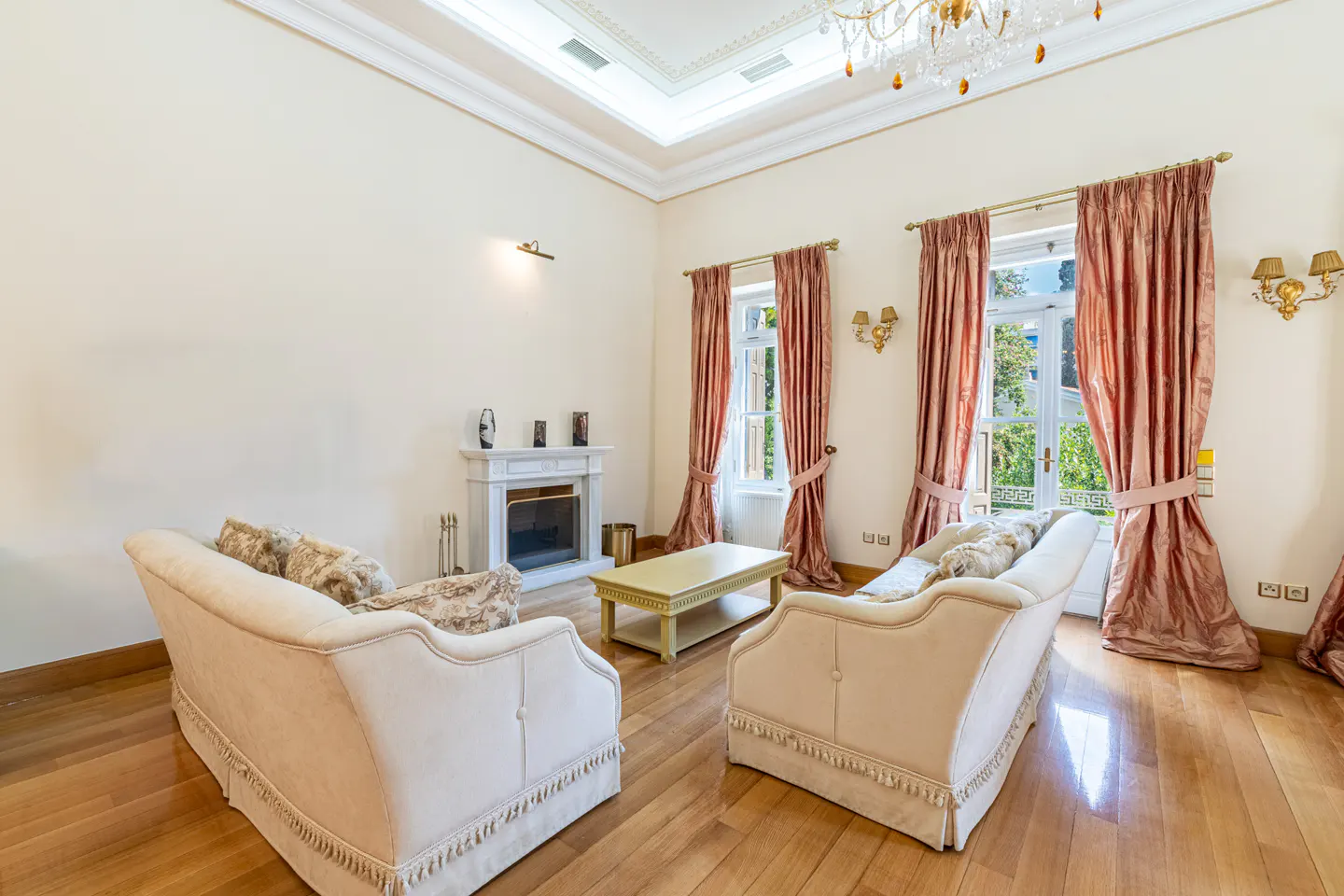 Bright living room with hardwood floors, two sofas, a fireplace, and pink curtains. A chandelier hangs from the ceiling.