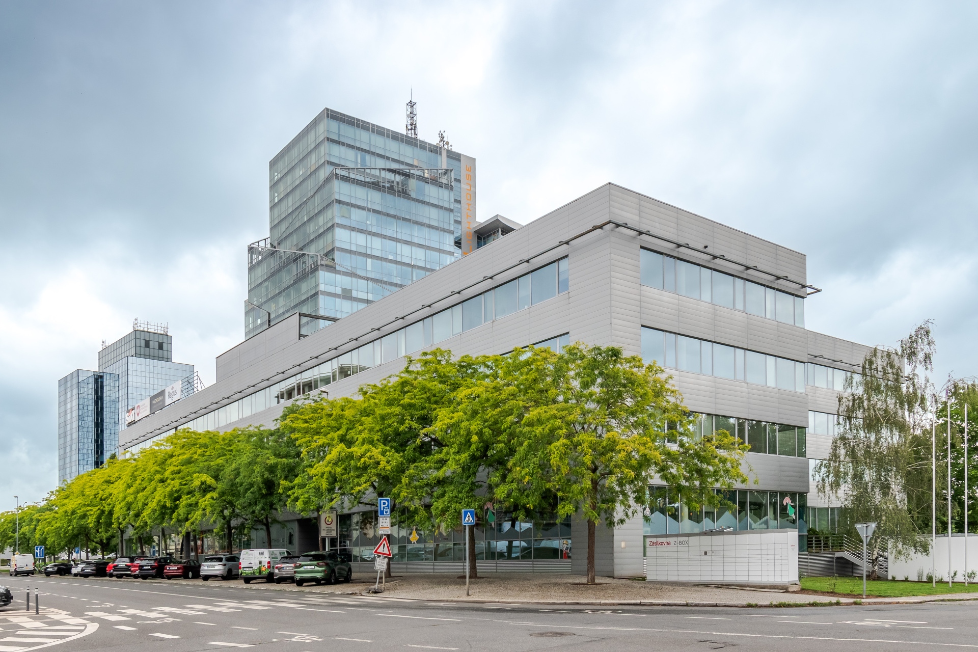 Modern gray office building with glass facade, surrounded by green trees, viewed from a street corner under a cloudy sky.