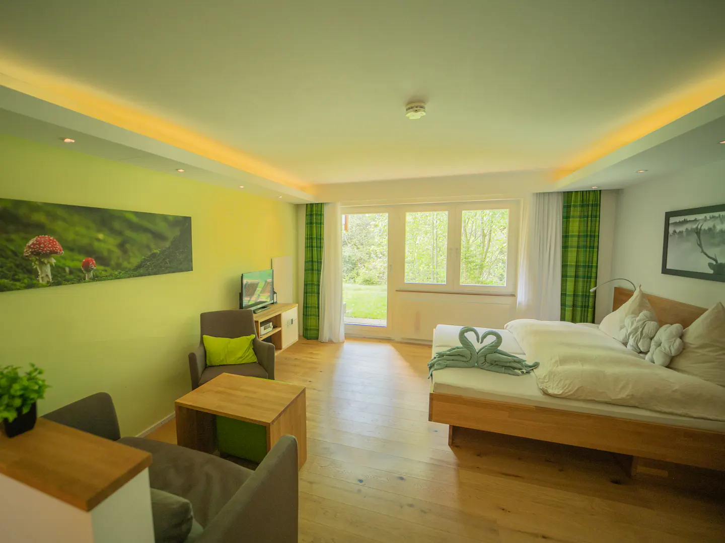 Bright hotel room with a bed, chairs, and a view of greenery outside the window. Towel swans sit on the bed. Mushroom art hangs on the wall.