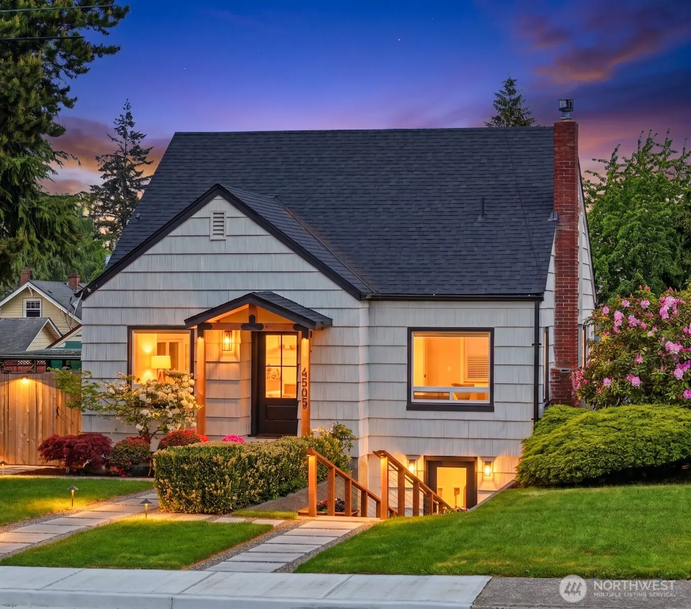 Exterior of a white, one-story house with a dark roof, brick chimney, and manicured lawn at dusk.