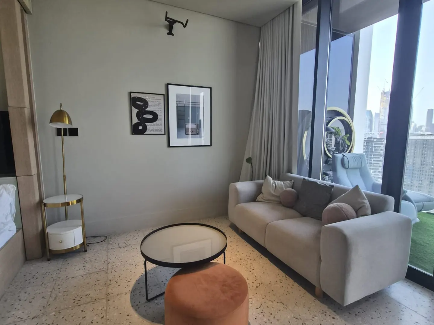 Modern living room with a beige sofa, round coffee table, orange ottoman, gold floor lamp, and city view through windows.