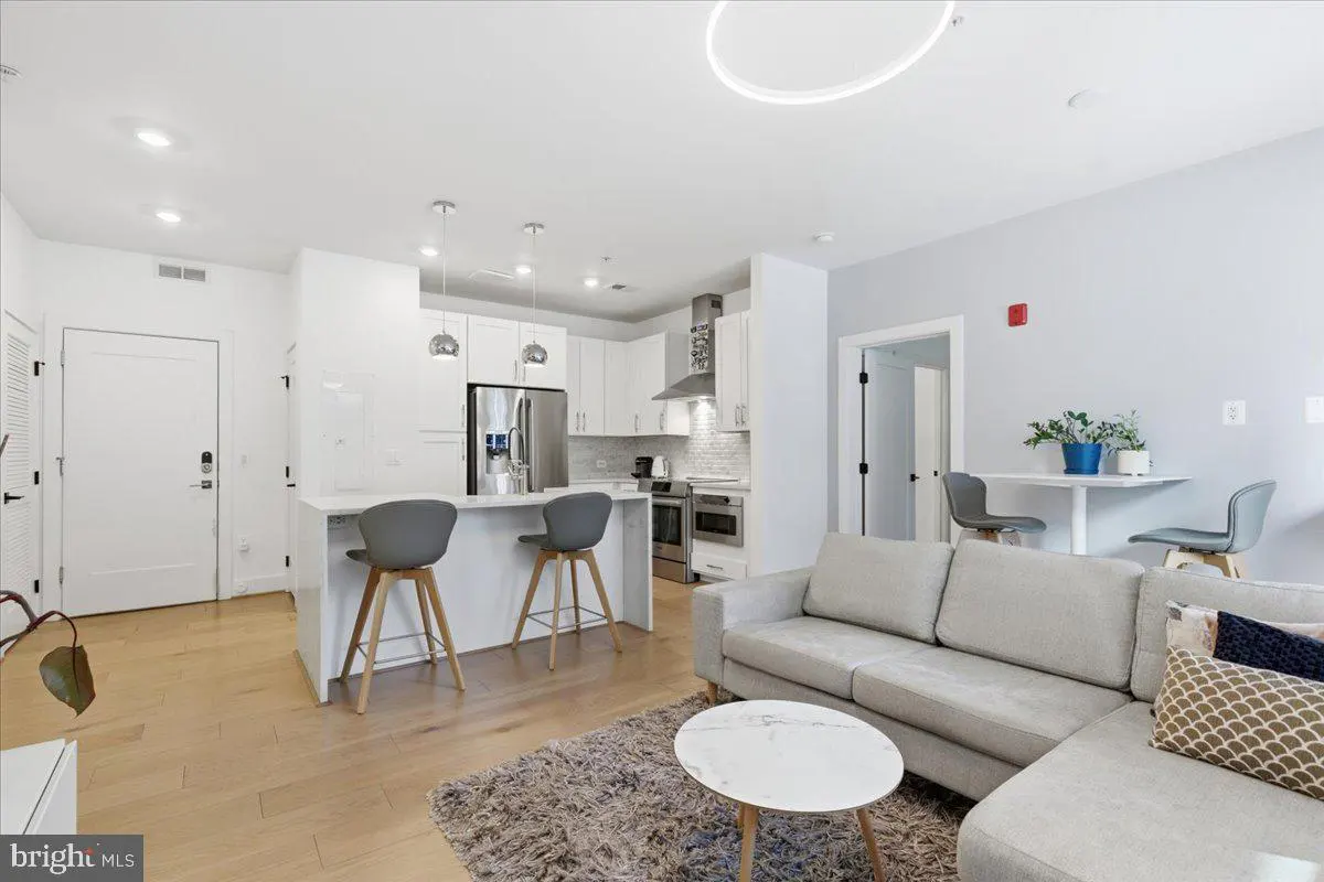 Bright, open-concept living space with white kitchen, gray stools, and a light gray sectional sofa. Hardwood floors and modern lighting.
