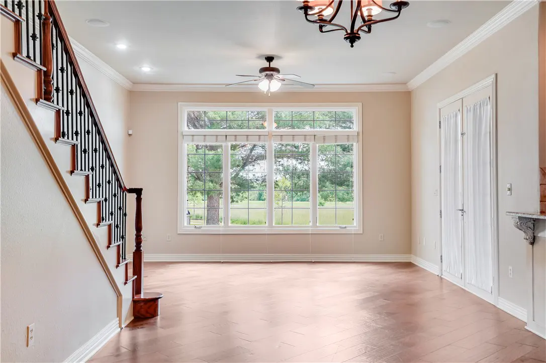 Bright, empty living room with wood floors, a large window overlooking a green yard, and a staircase on the left.