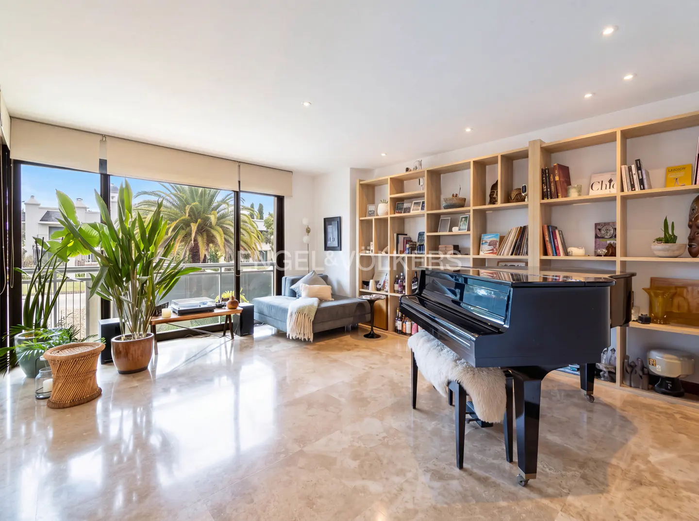 Bright living room with marble floors, a black grand piano, and a gray sofa. Large windows show palm trees. Built-in shelves line the wall.