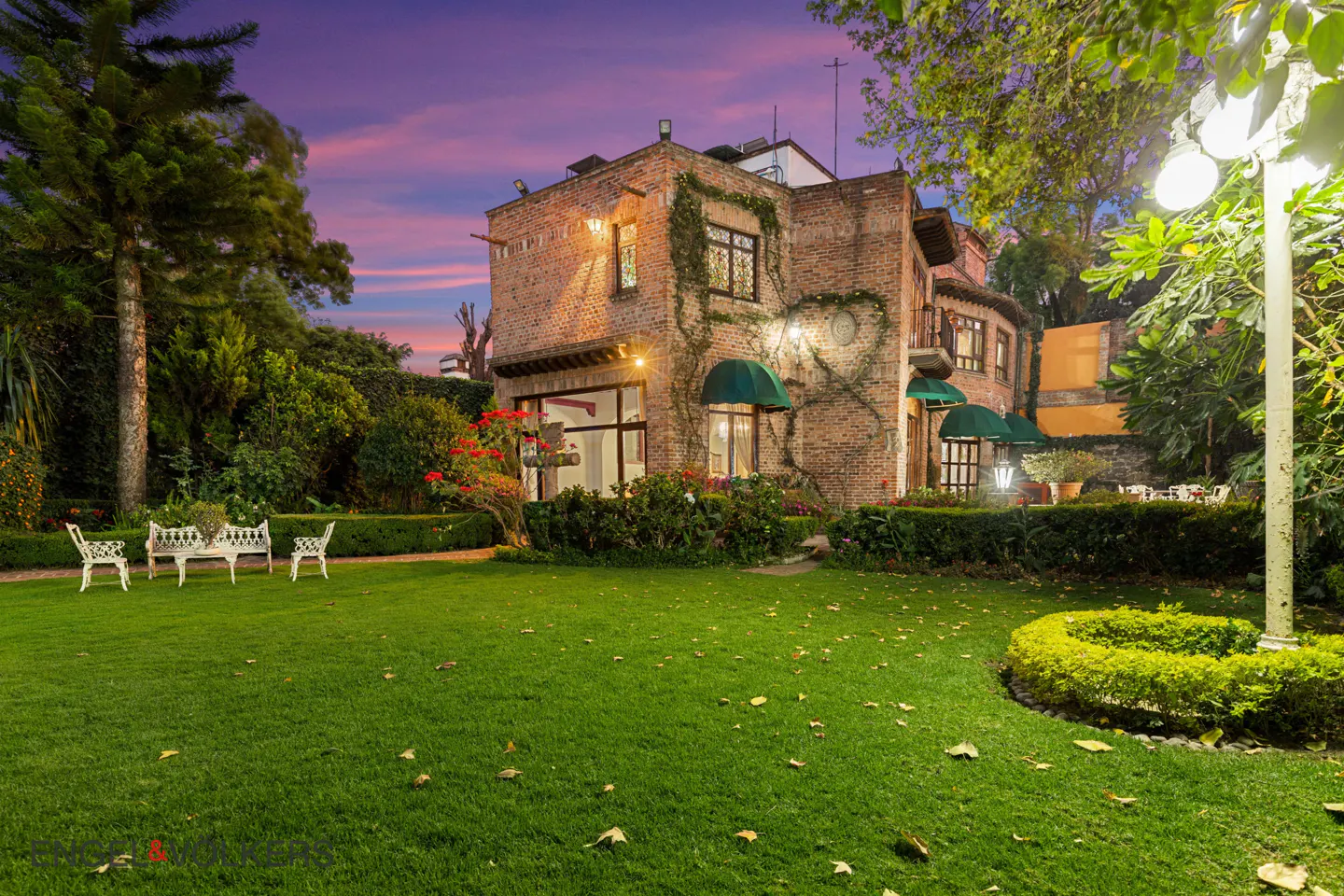 Brick house with green awnings and ivy, viewed from a lush green lawn with white garden furniture at dusk.