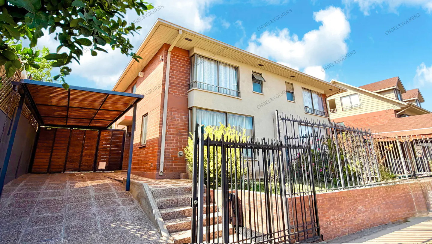 Two-story house with brick and stucco exterior, black iron fence, and covered parking area on a sunny day.