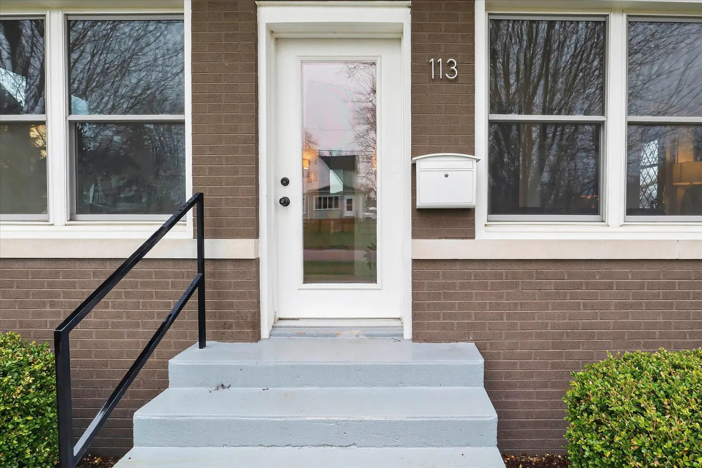 Exterior view of a brown brick house with a white door, mailbox, and trim. Three gray steps lead to the door, with a black handrail on the left. Address number 113 is above the mailbox.
