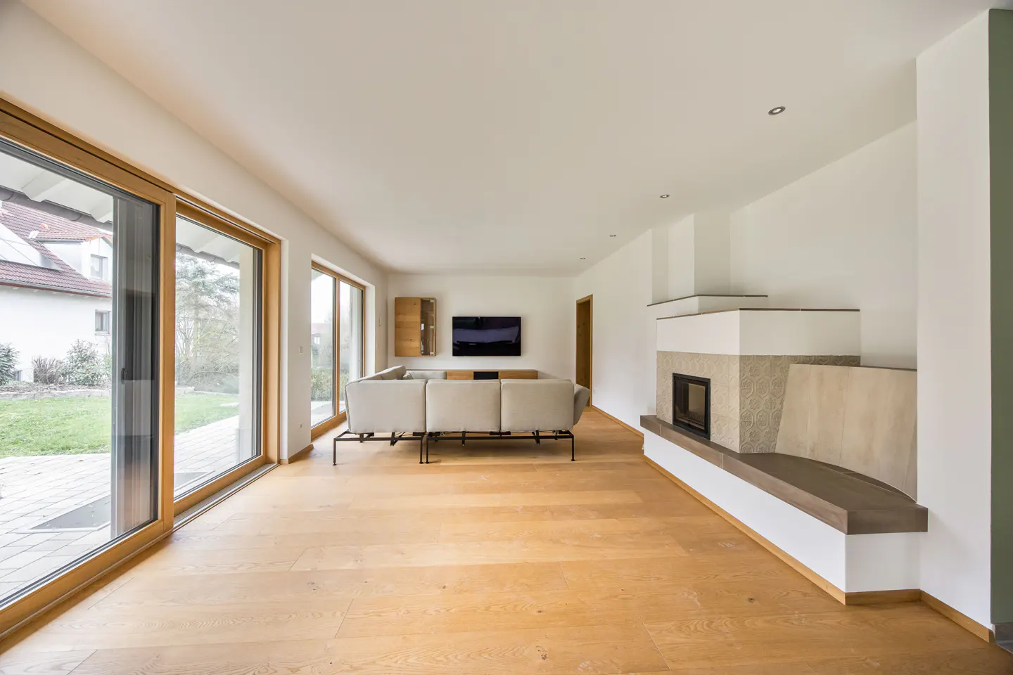 Bright, modern living room with wood floors, large windows, and a stone fireplace. A gray sofa faces a wall-mounted TV.
