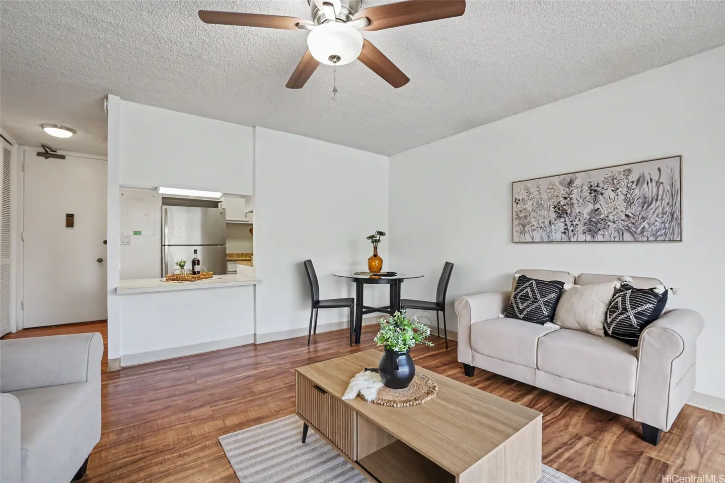 Living room with white walls, wood floors, and a ceiling fan. A sofa, coffee table, and dining table furnish the space. Artwork hangs on the wall.