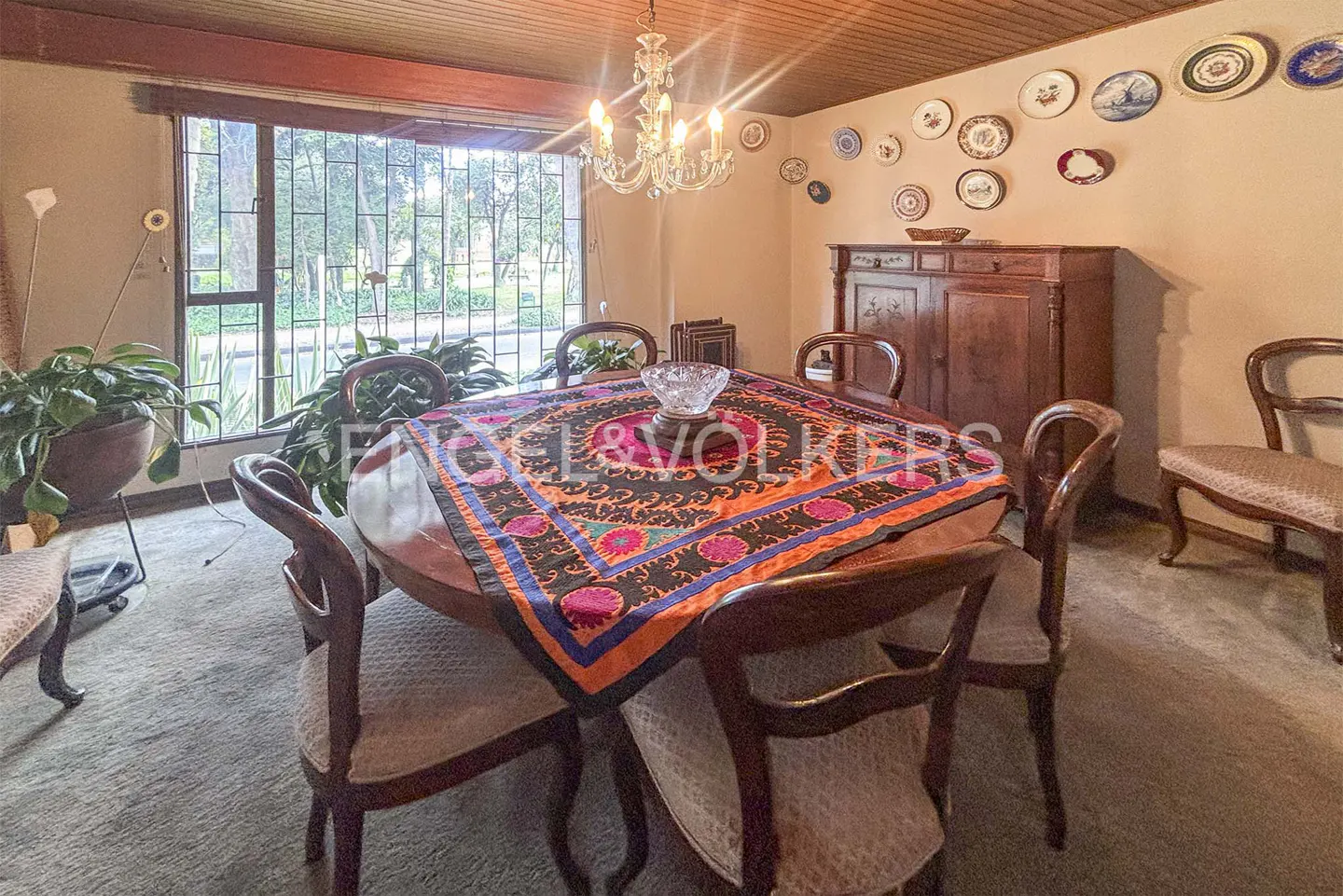 A dining room with a wooden table covered by a colorful cloth, surrounded by wooden chairs. A chandelier hangs above, and decorative plates adorn the wall.