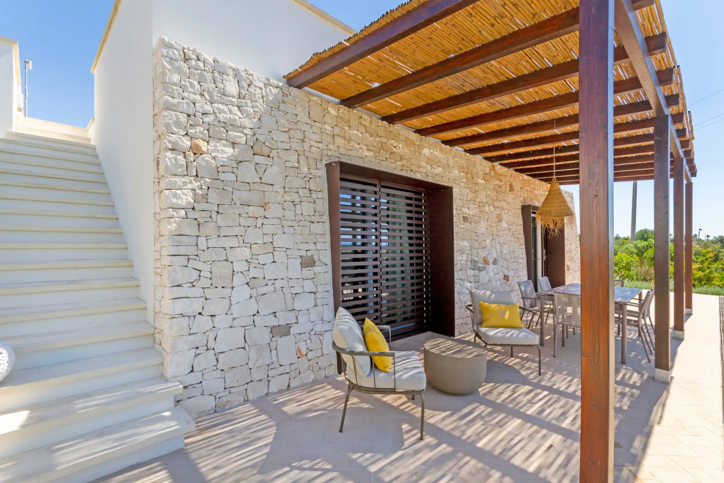 Patio with stone wall, wooden pergola, outdoor seating, and stairs leading to a white building under a bright blue sky.
