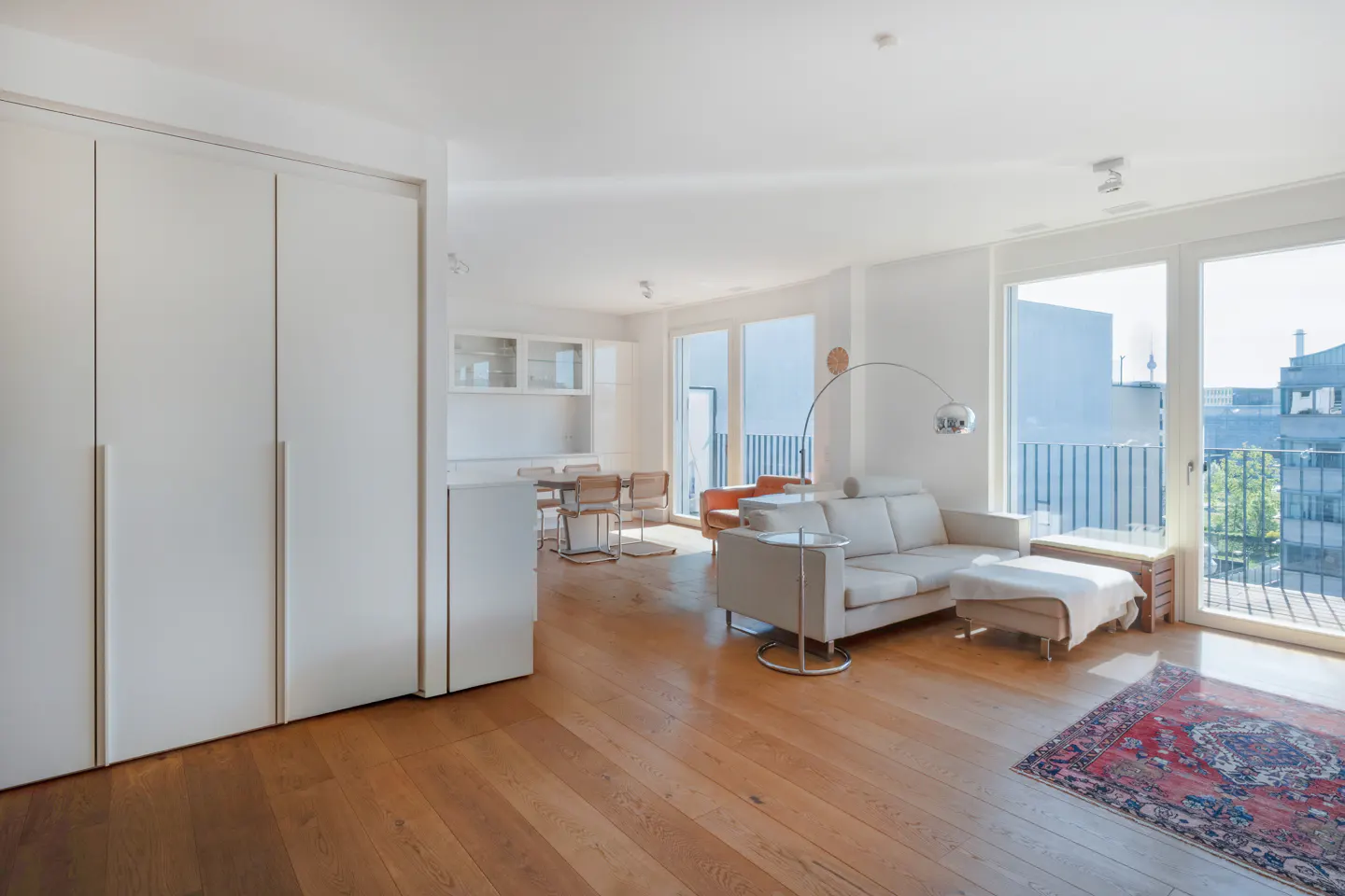 Bright, modern living room with hardwood floors, white walls, a beige sofa, and a balcony overlooking a city skyline.