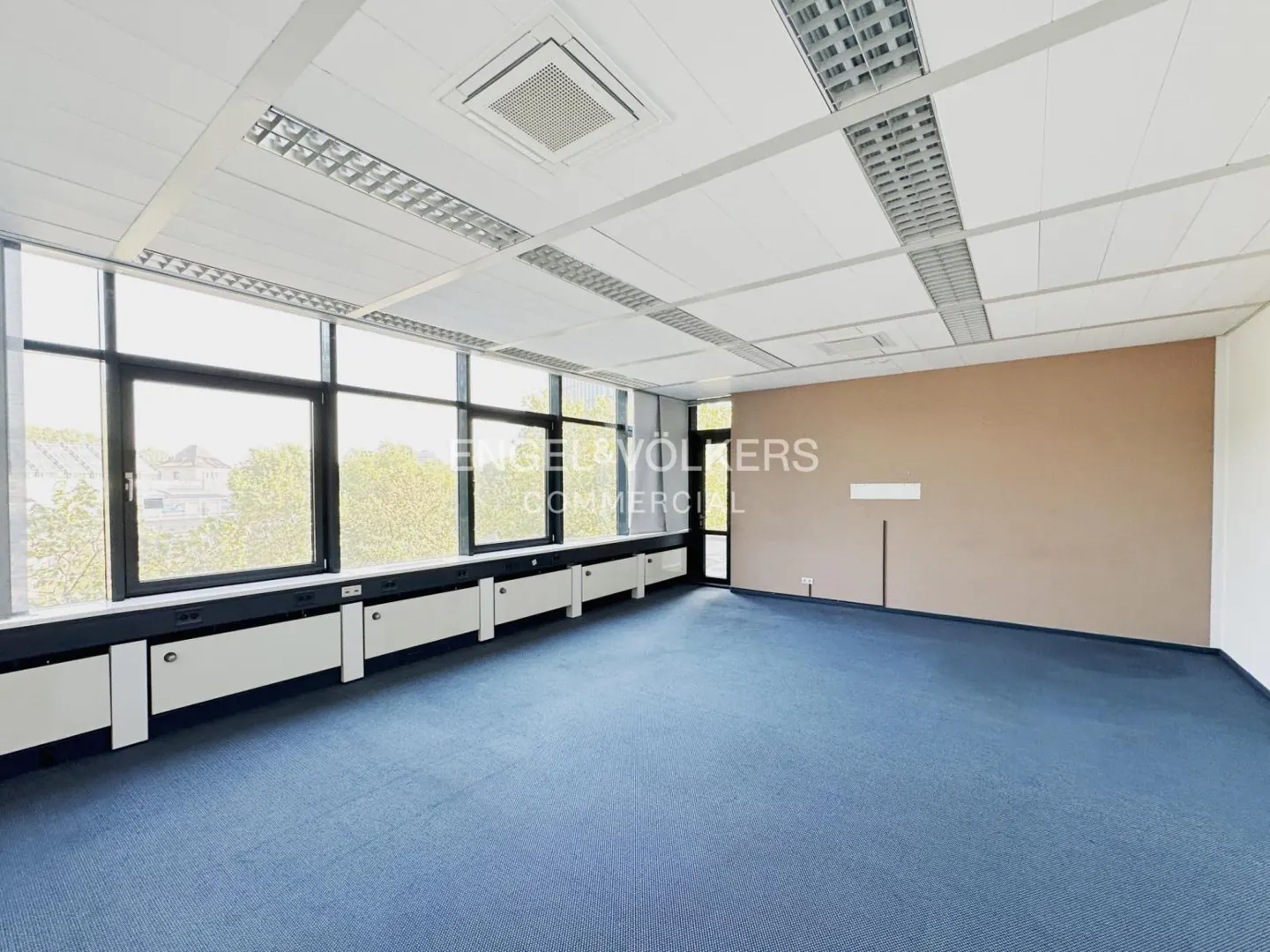 Empty office space with blue carpet, beige wall, and large windows. White ceiling with recessed lighting. Engel & Völkers logo visible.