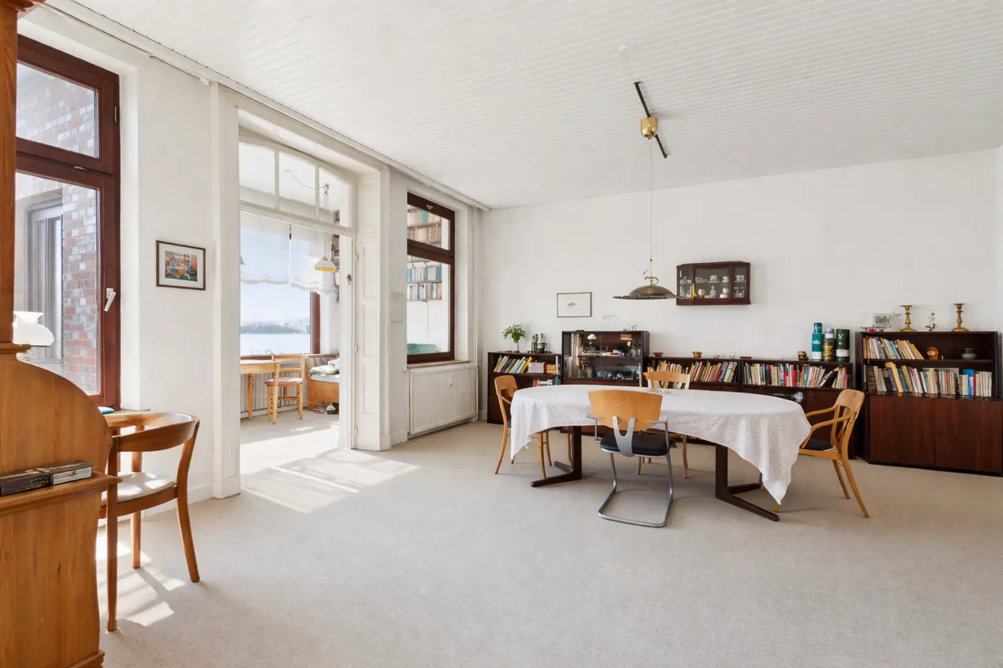 Bright, airy dining room with white walls, light carpet, and a large oval table with chairs. Bookshelves line the back wall. A view of the water is visible through an open doorway.