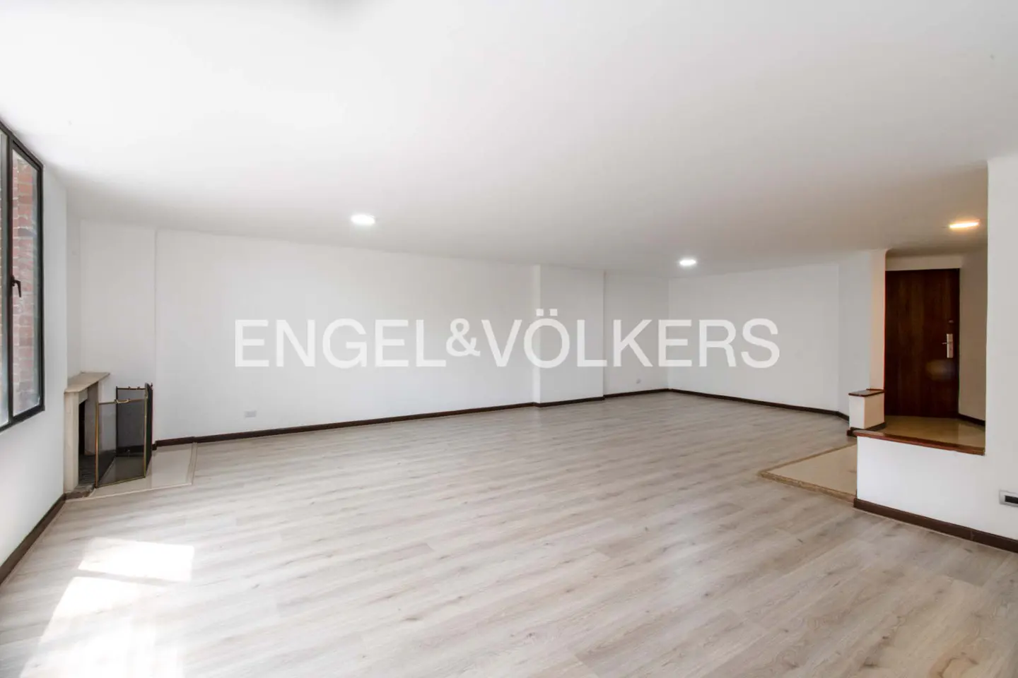 Spacious, empty living room with white walls, light wood-look flooring, a small fireplace, and a dark wooden door.