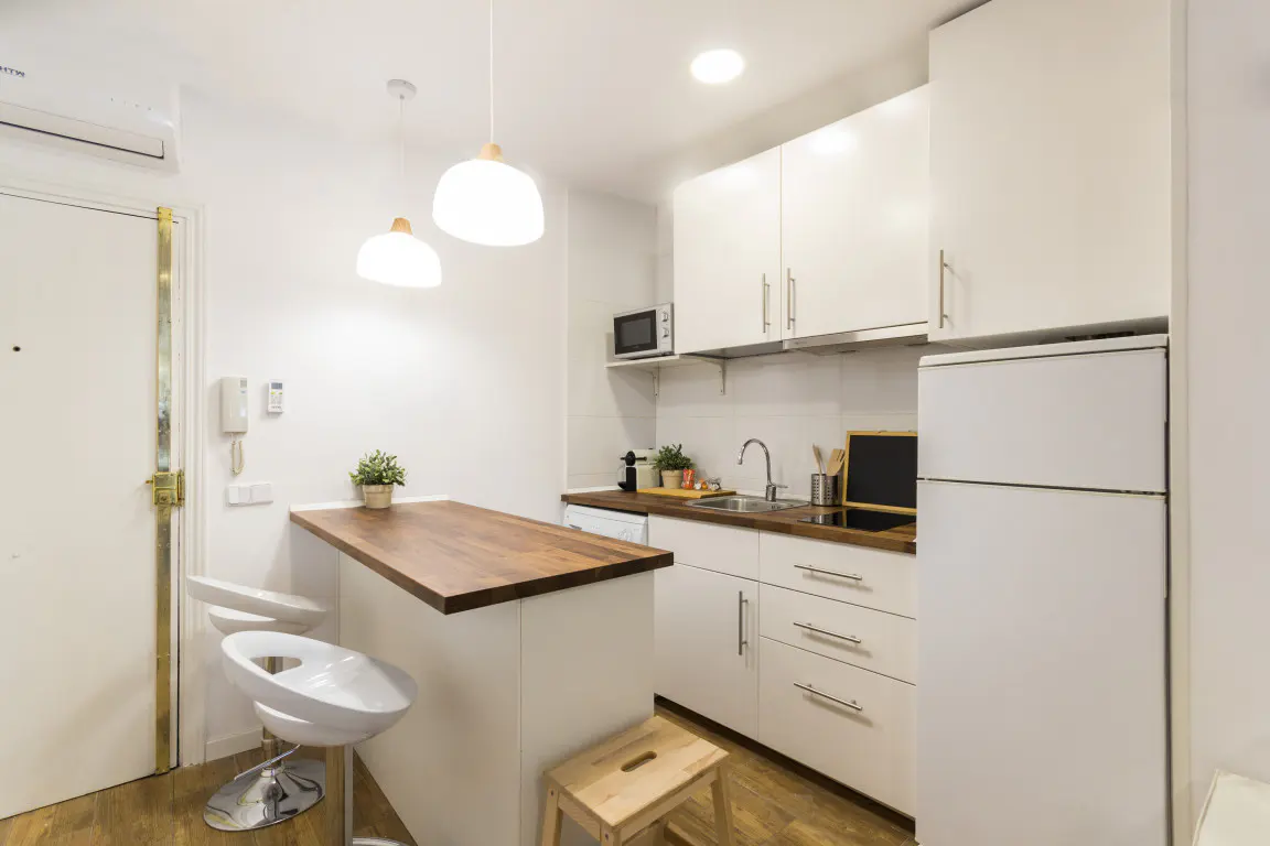 Bright, compact kitchen with white cabinets, wood countertop island, and two white stools. Appliances include a microwave and refrigerator.