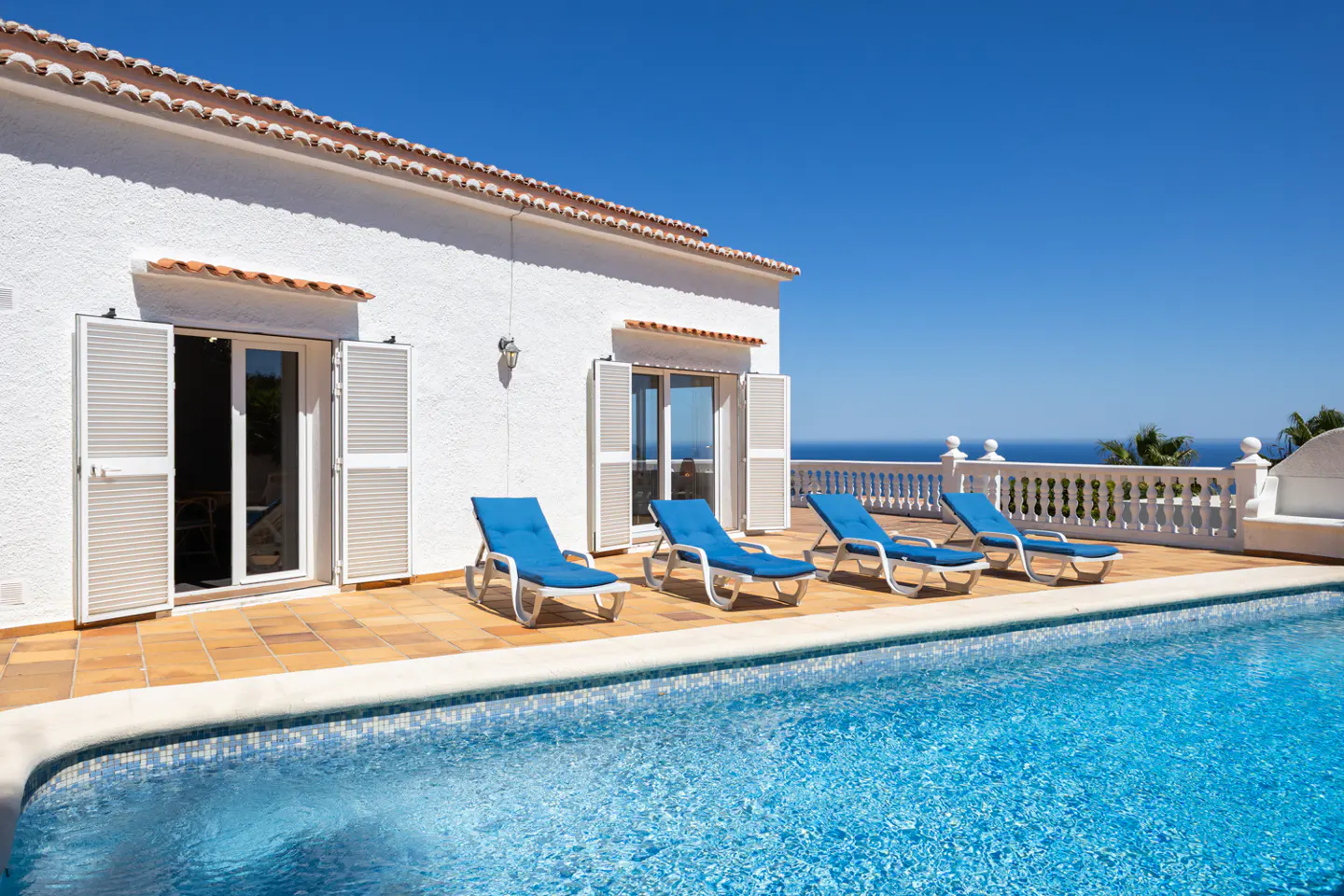 A white house with a pool and four blue lounge chairs on a sunny day.