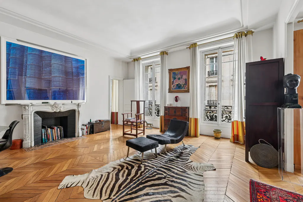 Bright living room with white walls, herringbone wood floors, and a zebra-print rug. A fireplace with blue art above it, and windows with white curtains.
