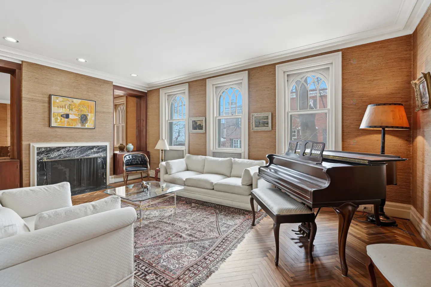 Living room with white sofas, a grand piano, and a fireplace. Three arched windows let in natural light. The walls are textured in a warm, neutral tone.