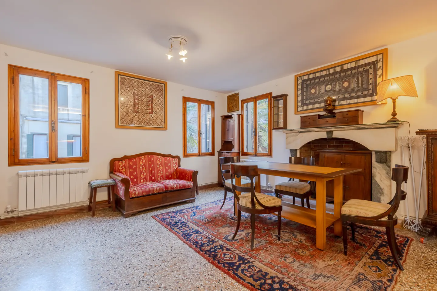 A vintage living and dining room with a red patterned sofa, wooden dining set on a large rug, and a stone fireplace.