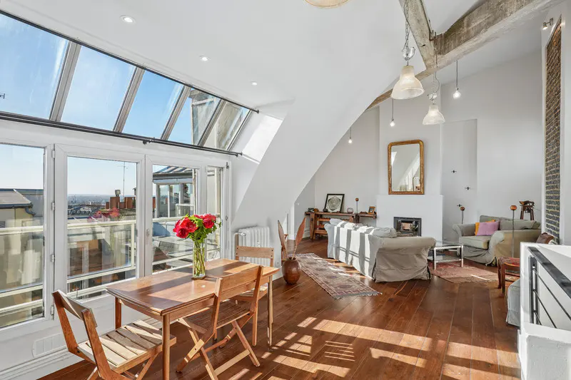 Bright, airy apartment interior with wood floors, white walls, and a skylight. A table with red roses sits near a window overlooking the city.