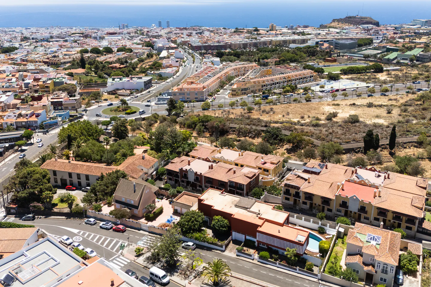 Aerial view of a sunny residential area with terracotta-roofed houses, roads, and distant ocean views.