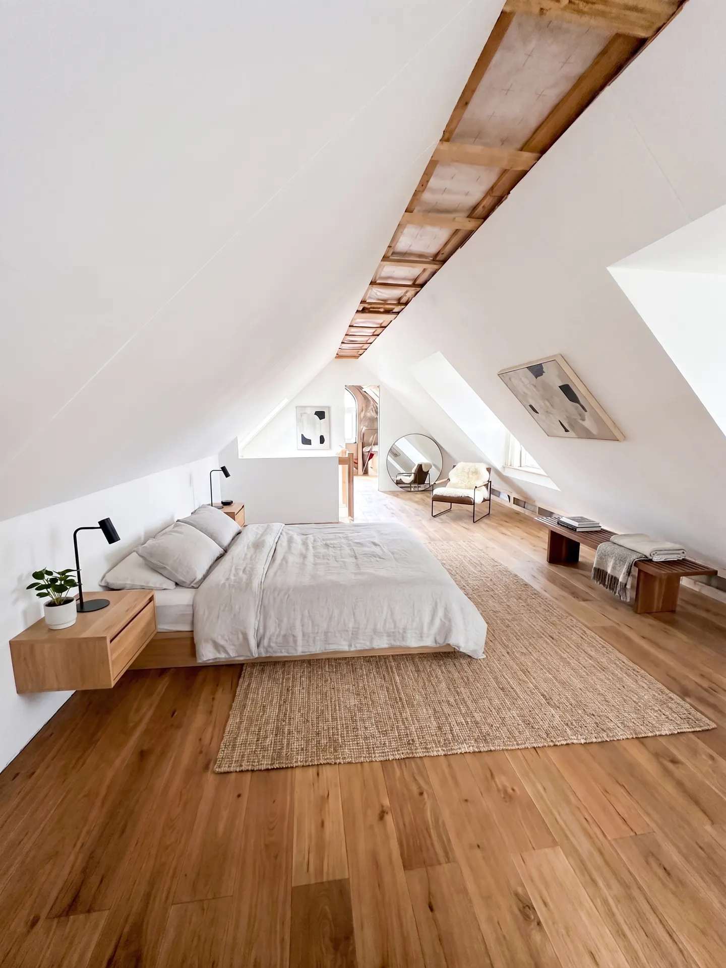 Attic bedroom with wood floors, white walls, and exposed ceiling beams. A platform bed with light gray linens sits on a jute rug. A chair and bench are in the background.