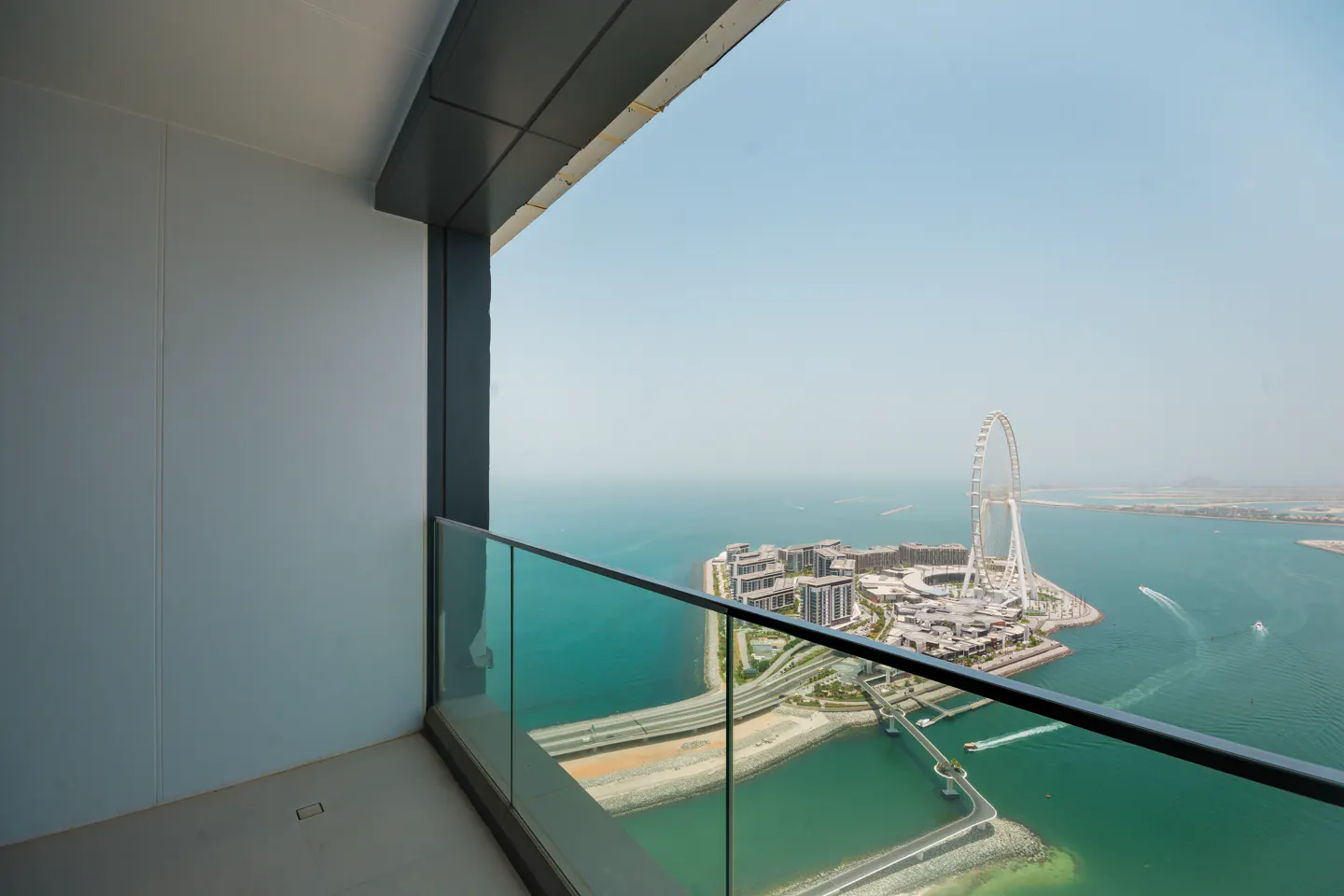 View from a high-rise balcony with glass railings overlooking a city island with a large Ferris wheel on the turquoise sea.