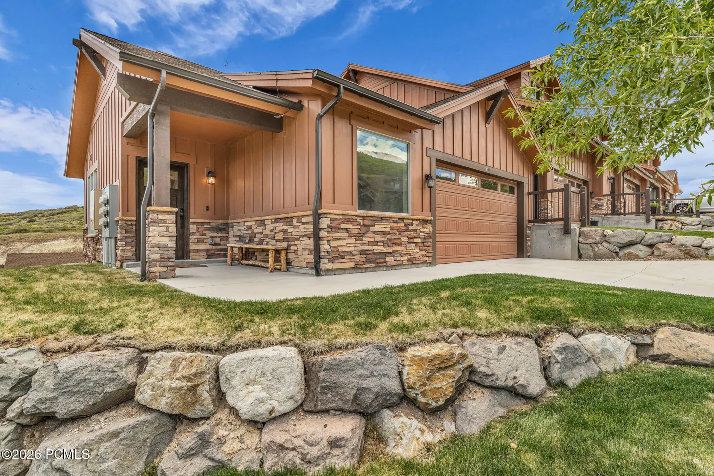 Modern mountain home with brown vertical siding, stone accents, a garage, and a front porch with a bench under a blue sky.