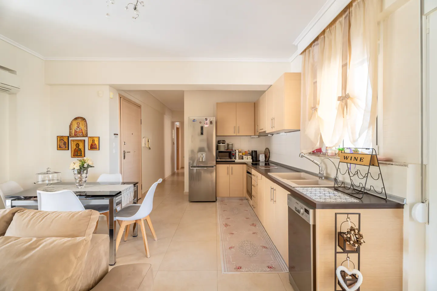 Bright apartment interior with beige cabinets, stainless steel appliances, dining table, and tan sofa. Icons hang on the wall.