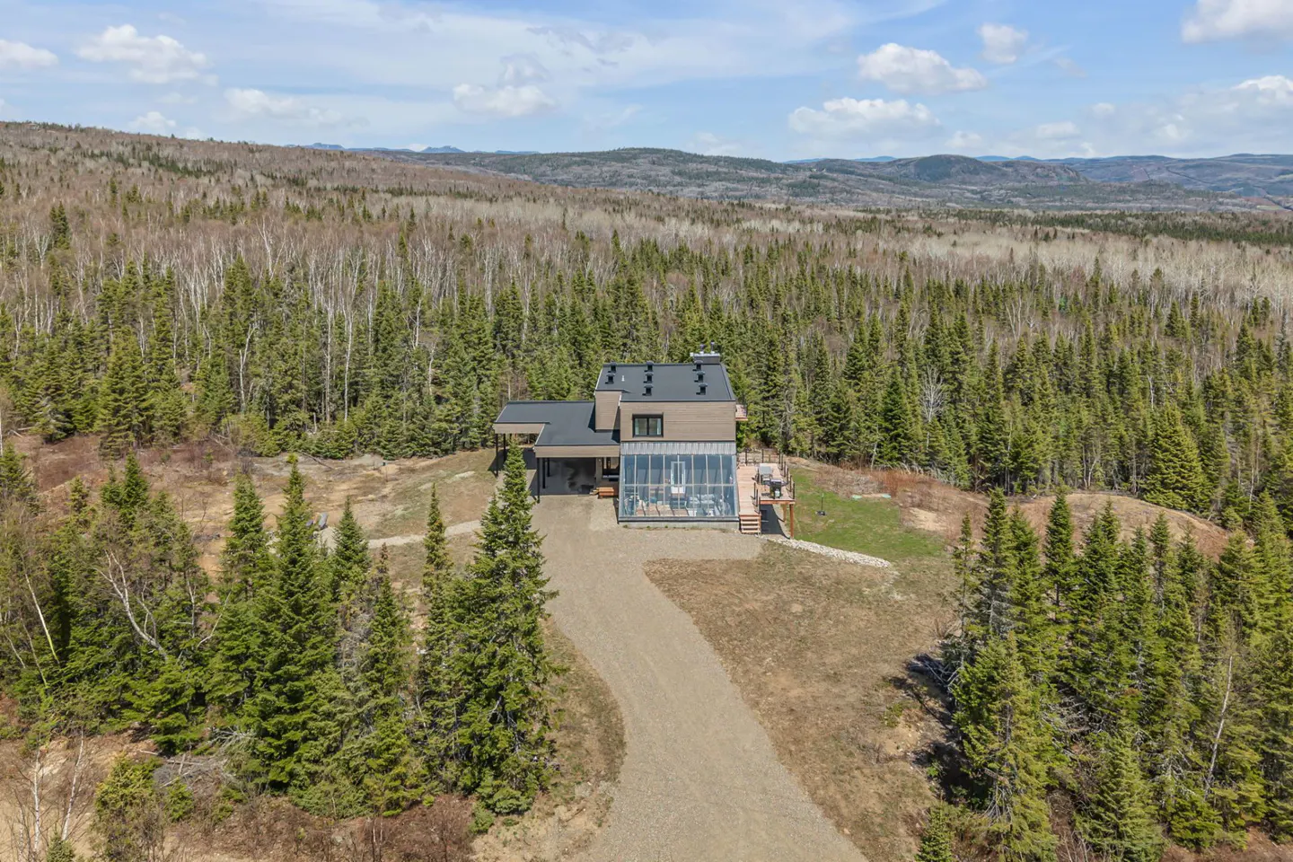 Modern home with glass sunroom on a hill surrounded by a forest. The house has a gravel driveway and a deck.