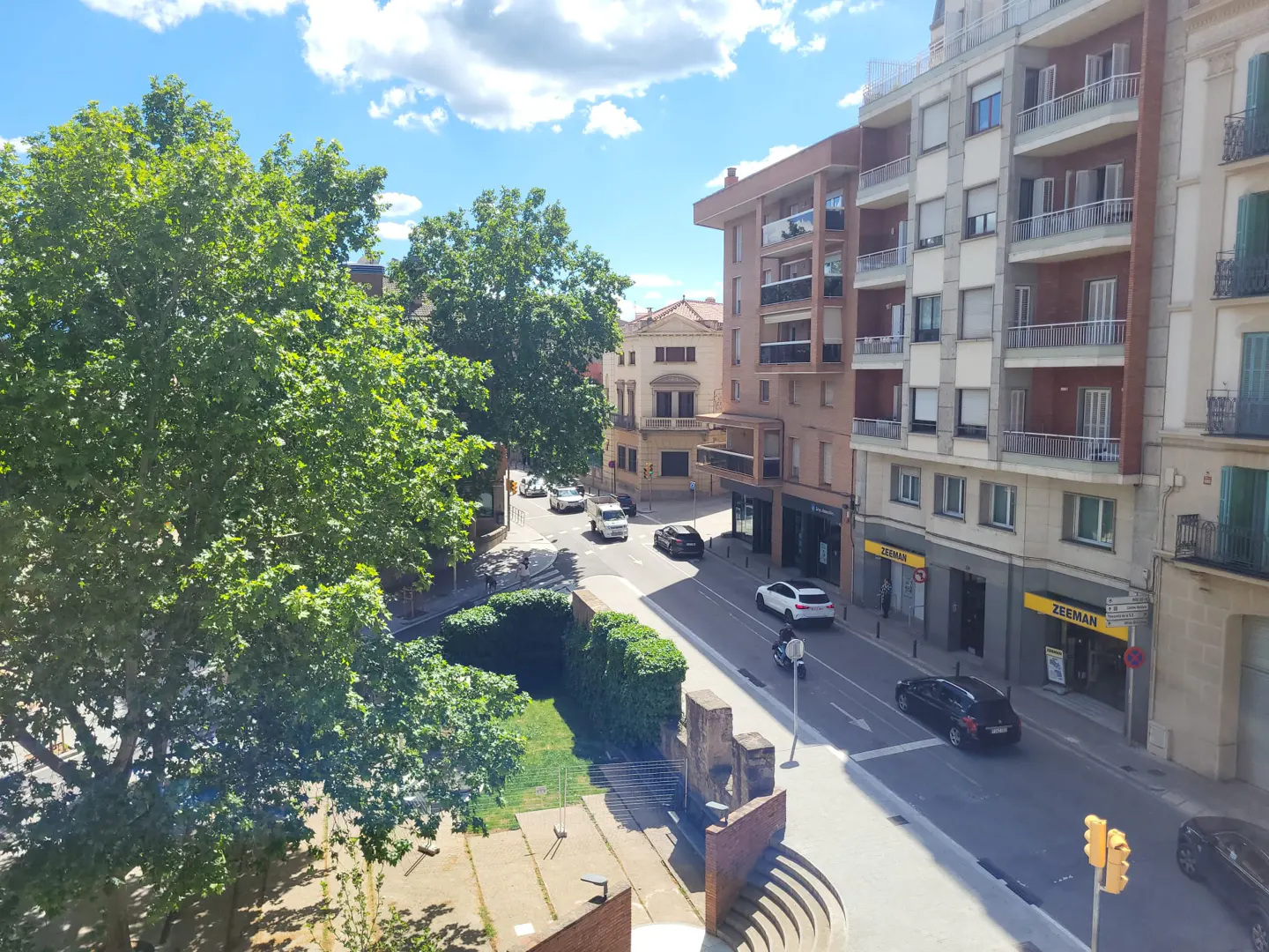 Street view of European city with cars, buildings, trees, and blue sky with clouds. A Zeeman store is visible on the right.
