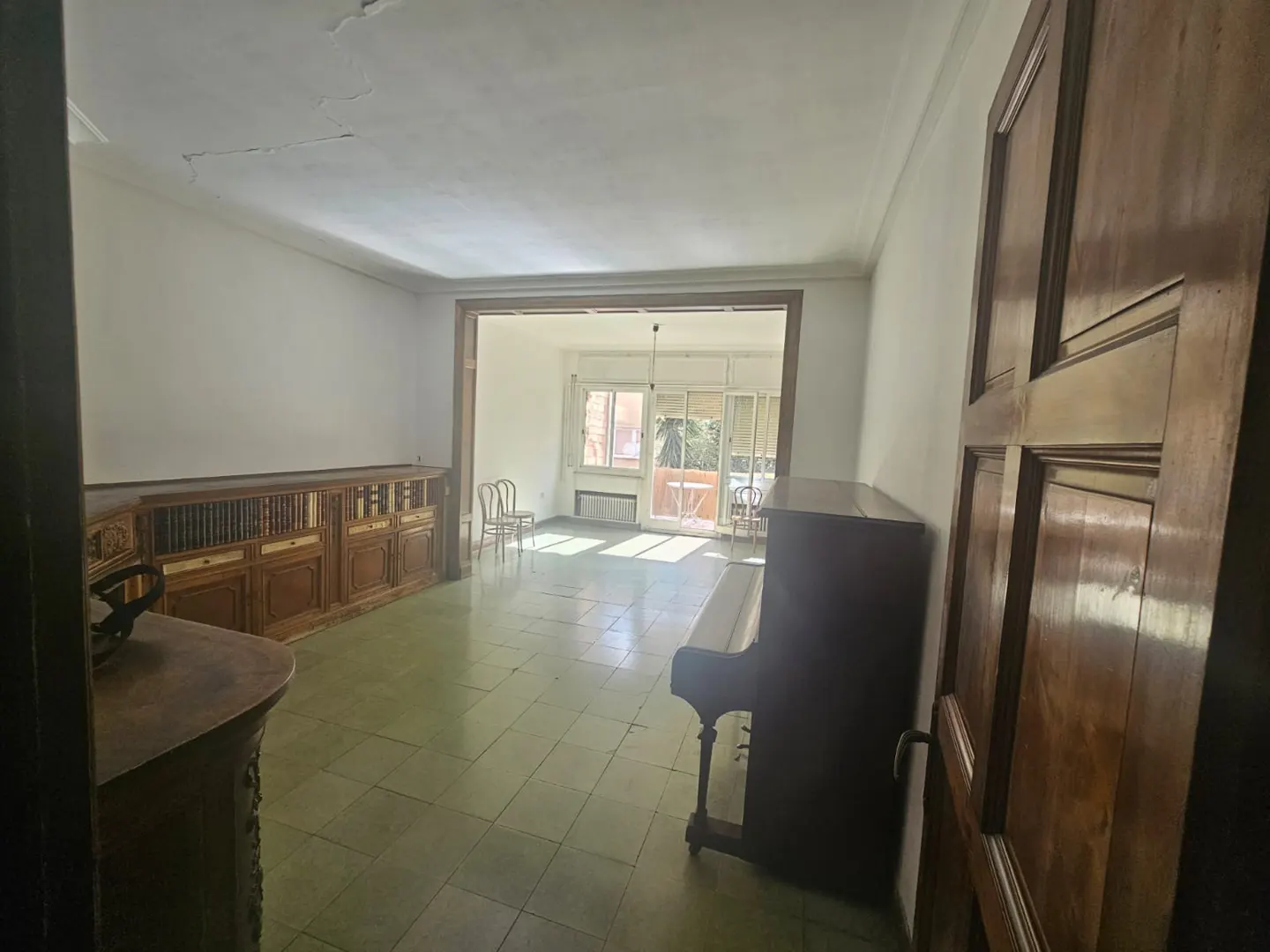 Spacious living room with light green tiled floors, a wooden bookshelf, an upright piano, and a bright sunlit balcony.